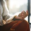 A close-up of a person meditating, wearing neutral-toned clothing, bathed in soft morning sunlight. The hand in a mudra gesture conveys calm and mindfulness.