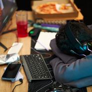 Young adult Black man wearing headphones sleeping at desk with head resting on arms near computer monitor, keyboard, smartphone, game controller, pizza box and drink visible on table