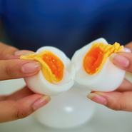 A close-up view of two hands gently splitting a soft-boiled egg, revealing its golden, runny yolk. The image showcases the delicate texture of the egg, with the creamy yolk oozing out against the smooth, cooked egg white.